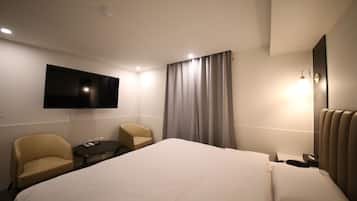 A hotel room with a large bed, two armchairs, a small table, a flat-screen TV, a bedside lamp, and a window with curtains.