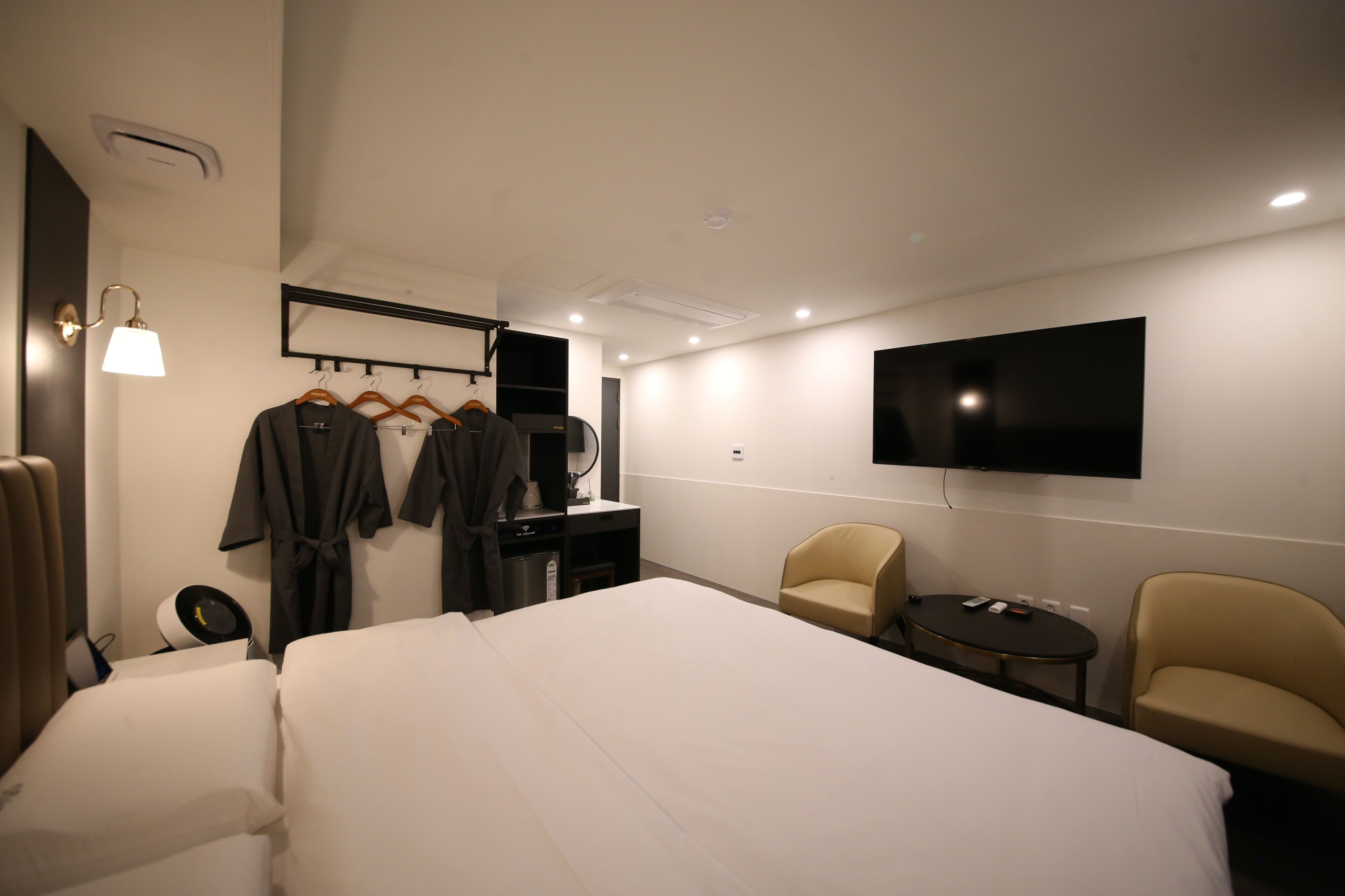 A hotel room with a large bed, a desk, a chair, a TV mounted on the wall, and a wardrobe with hanging clothes.