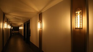 Un long couloir étroit, typique des hôtels, avec des lampes fixées aux murs et des portes de part et d’autre.