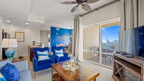 A modern living room with a blue accent wall, a ceiling fan, a large TV, and a balcony with a city view. - Luxury resort condo-10 th floor-Easy 4 min walk to beach, perfect time of year (Miramar Beach)
