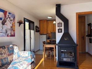 Una sala de estar con un sofá estampado, un estufa de leña negra, armarios de madera y una zona de cocina con refrigerador.