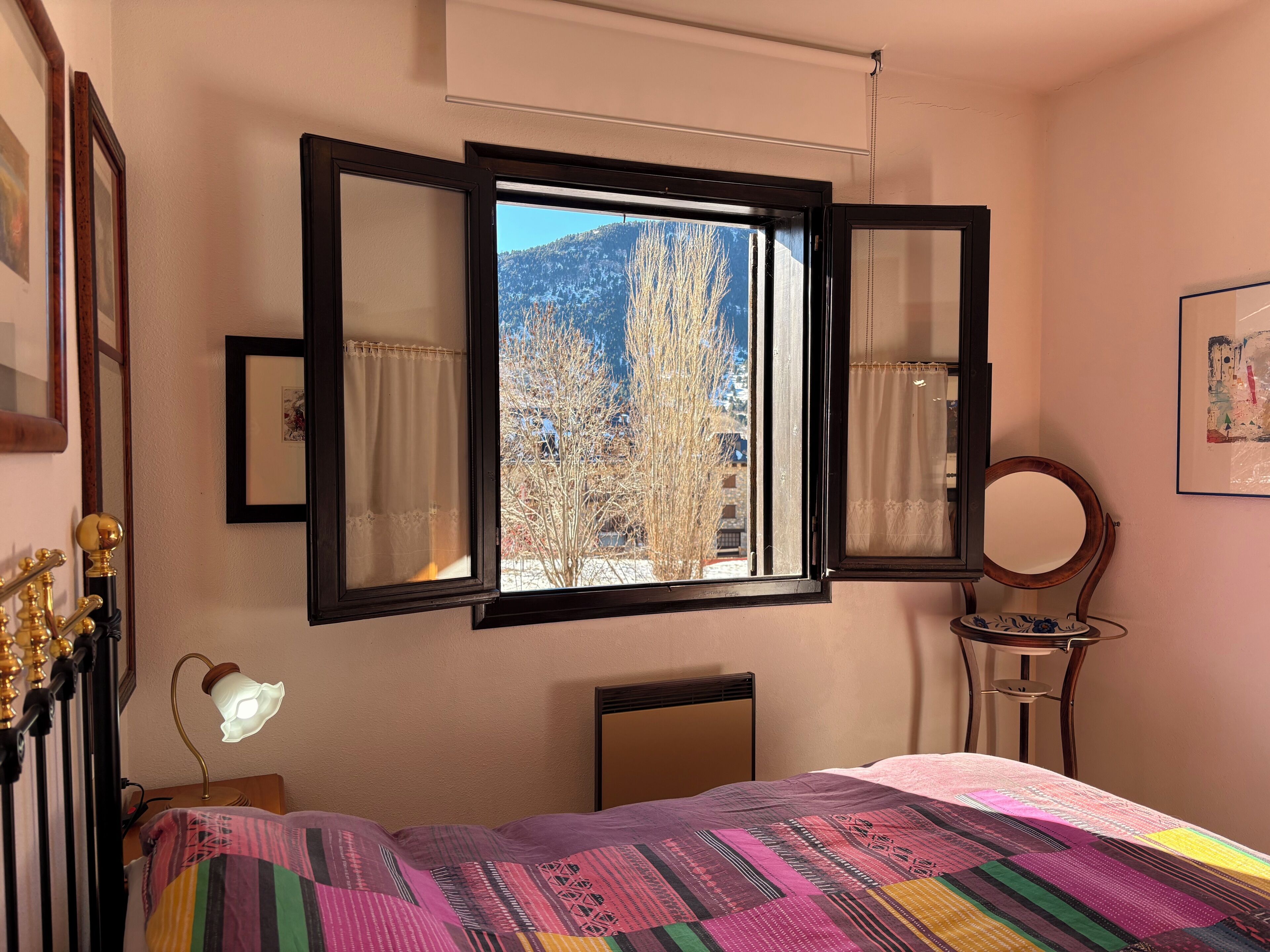 A bedroom with a bed, a window with curtains, a bedside lamp, a mirror, and a wall-mounted picture.