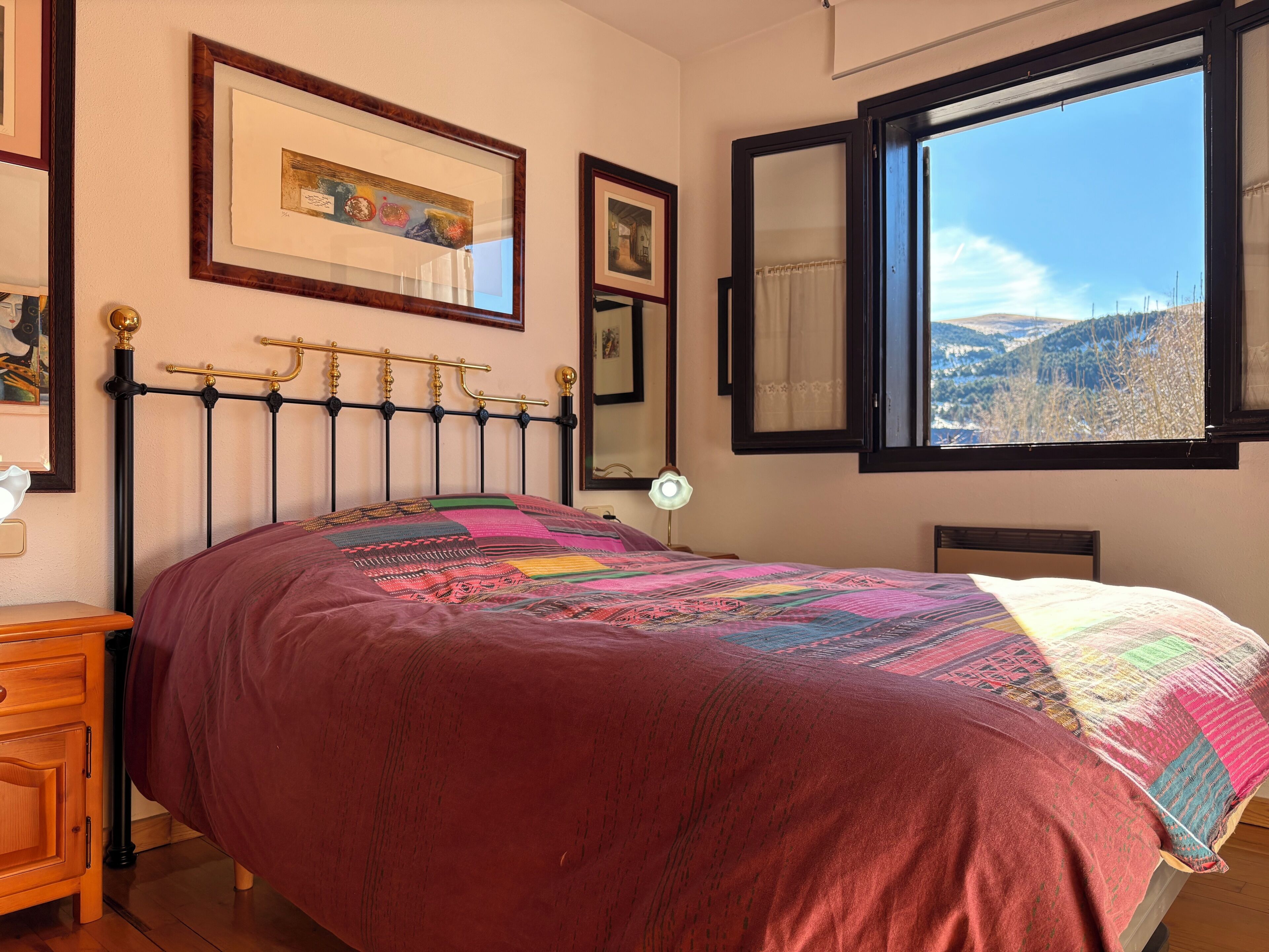 A bed with a patterned blanket, a bedside table, a lamp, and a window with a view of a landscape.