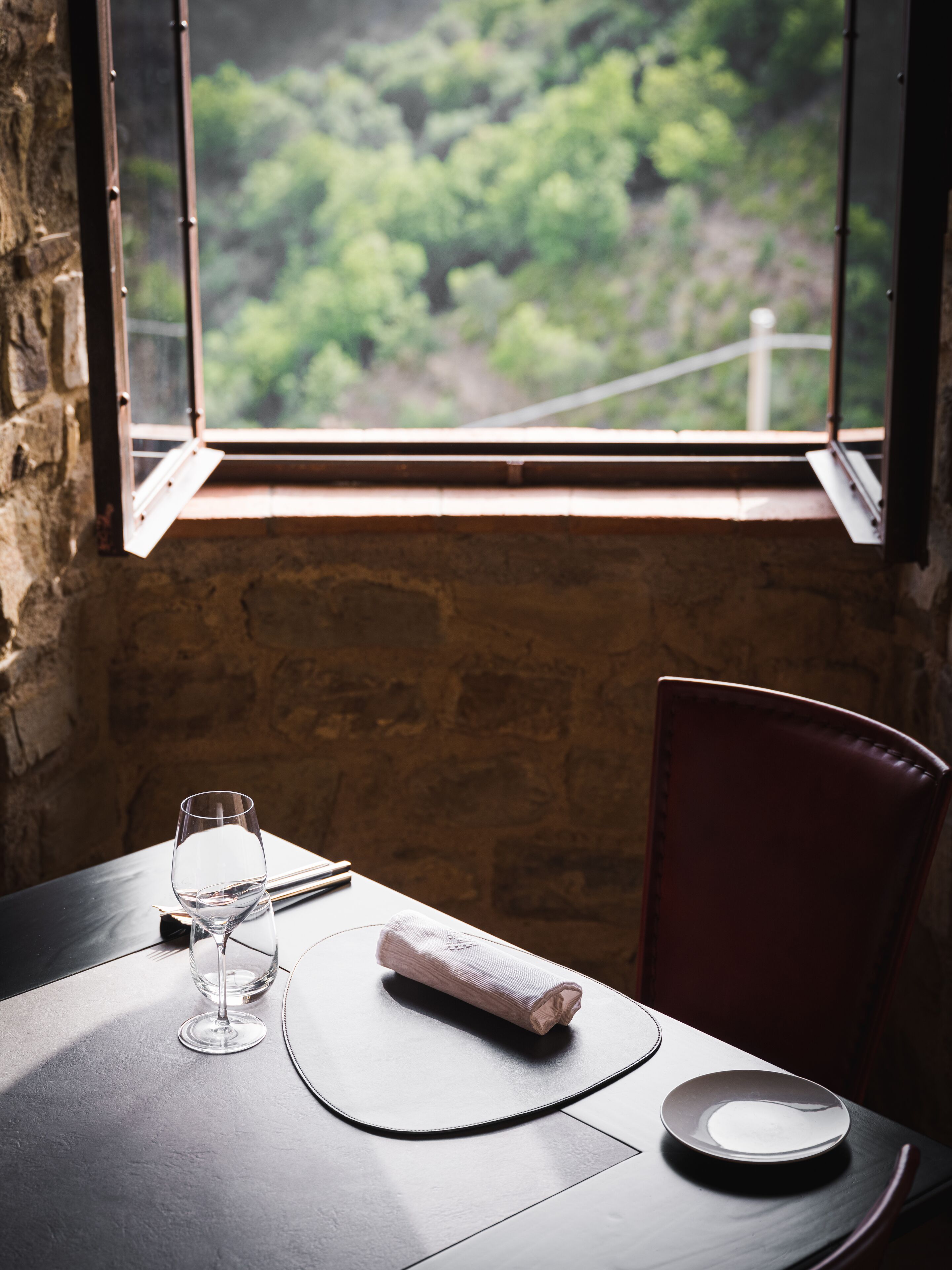 Un tavolo con un bicchiere di vino, un tovagliolo piegato e un piatto, sullo sfondo di un muro in mattoni con una finestra che si affaccia su una foresta.