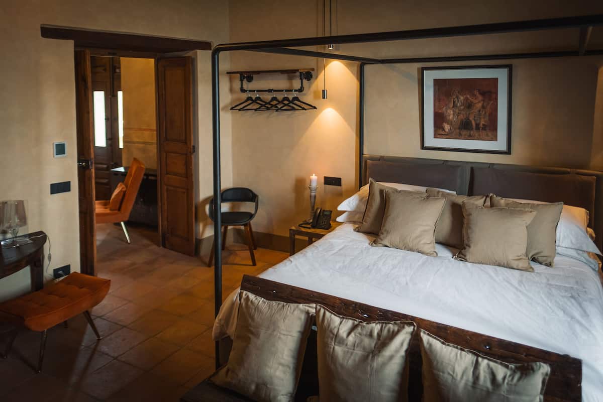 A hotel room with a four-poster bed, a bedside table with a lamp, a chair, and a coat rack.