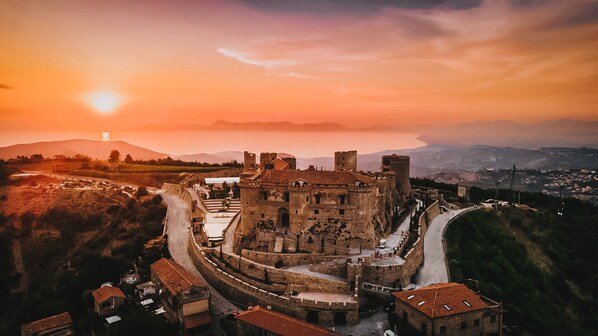 Un castillo histórico con un muro fortificado, un patio central y un paisaje circundante con montañas y un atardecer.