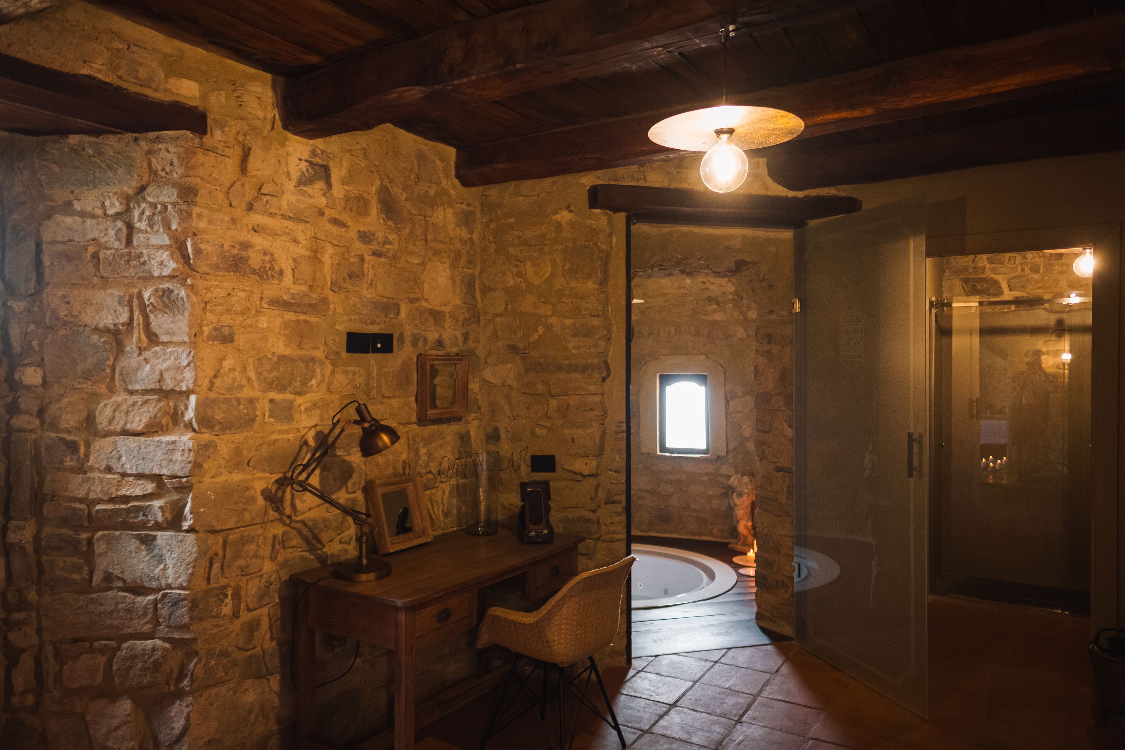 A stone-walled room with a wooden desk, a chair, a hanging light fixture, and a door leading to another room.