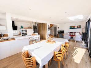 A modern interior with a dining area, kitchen, and living space, featuring a large island table and wooden chairs.