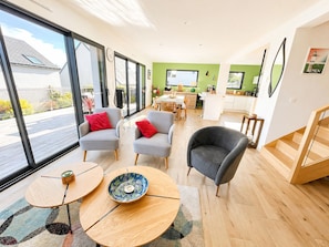 A modern living room with a dining area, large windows, and a kitchen in the background.