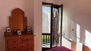 Une chambre avec une commode en bois, un lit recouvert d’une courtepointe rose et une porte-fenêtre donnant sur un balcon.