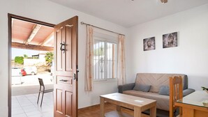 Una habitación con puerta de madera, ventana con cortinas, un sofá, una mesa y dos cuadros en la pared.