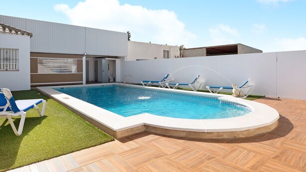 Una piscina con sillones de descanso, una zona cubierta y un edificio blanco.
