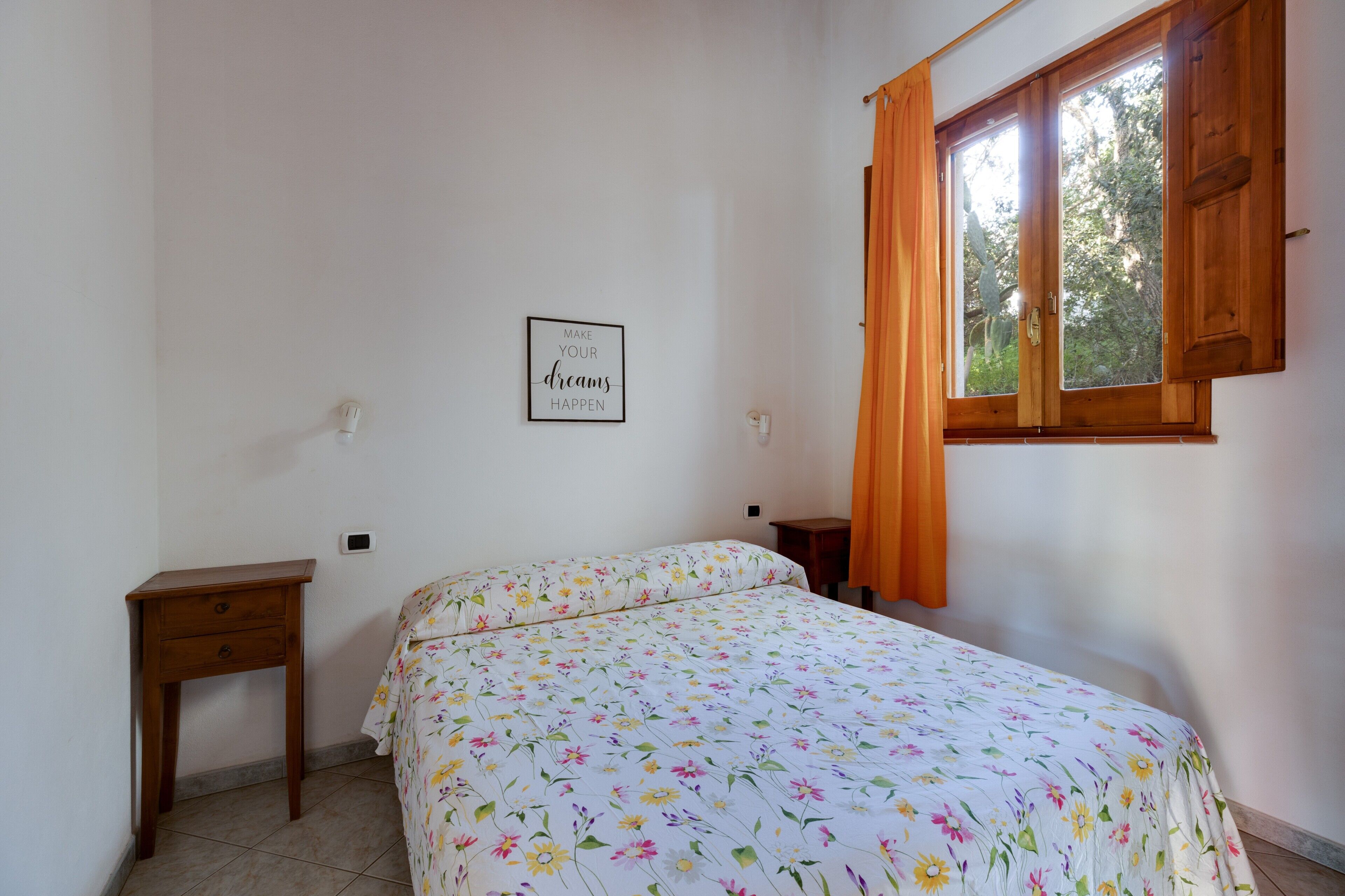 Une chambre à coucher avec un lit, une petite table de chevet, une lampe fixée au mur, une fenêtre avec des rideaux et une citation inspirante sur le mur.