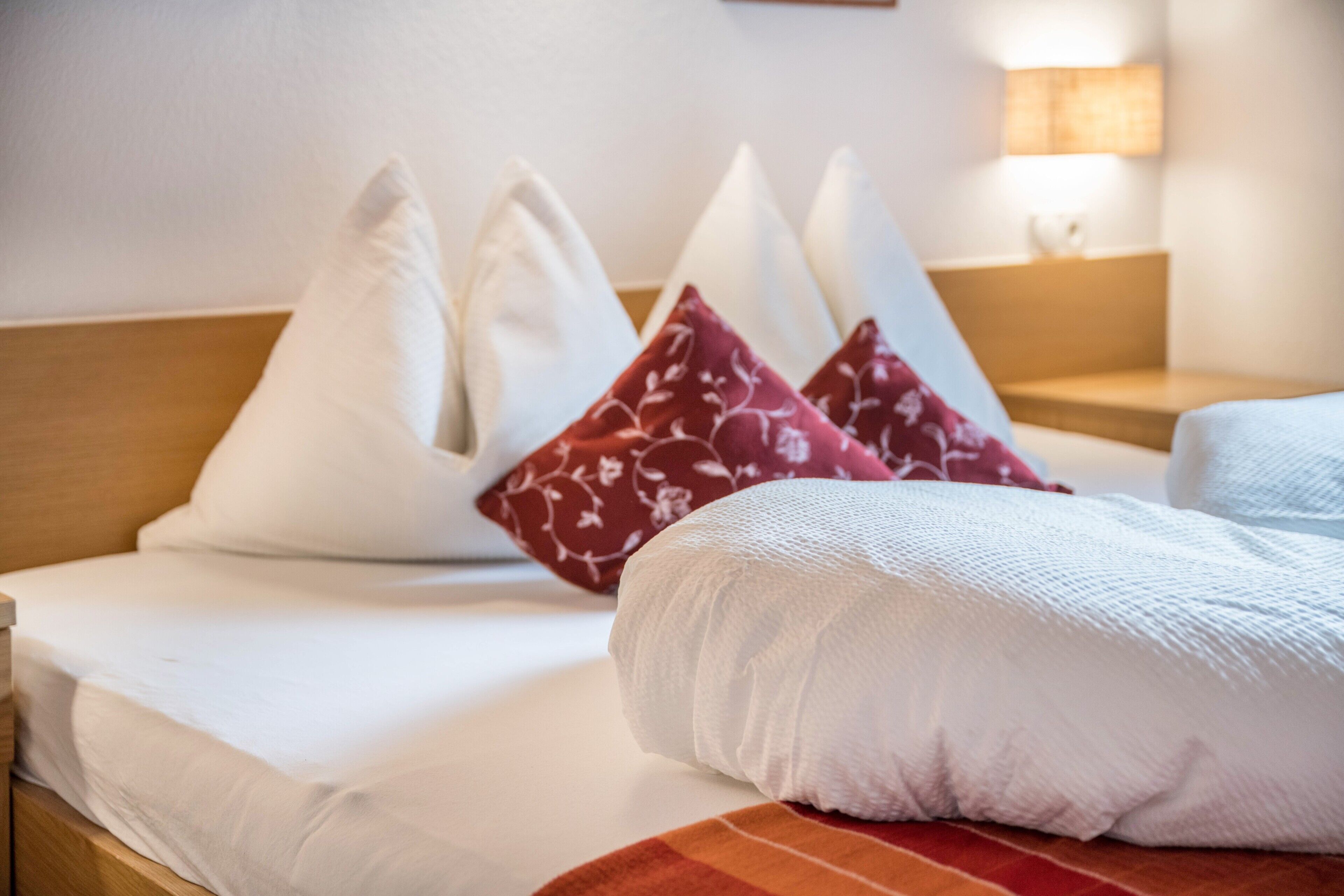 Ein Hotelzimmer mit einem ordentlich bezogenen Bett, Kissen in Weiß und Rot mit Muster, einem Holzkopfstück und einer Nachttischlampe.