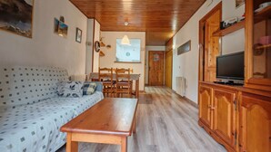 Een gezellige woonkamer met een houten plafond, een bank met gestreepte kussens, een houten tafel, een televisie op een houten kast, en een eethoek met stoelen en een tafel.