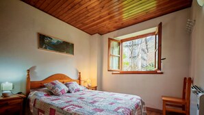 Een slaapkamer met een houten plafond, een bed met een sprei, een nachtkastje met een lamp, een stoel en een raam met uitzicht op bomen en gebouwen.