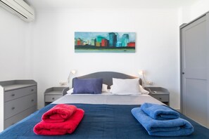 Une chambre d’hôtel comprenant un lit, deux tables de chevet, une petite table, un climatiseur fixé au mur et un tableau représentant un paysage urbain accroché au mur.