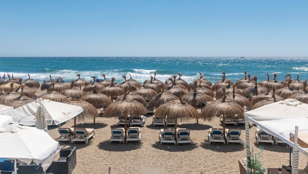 A beach with rows of sun loungers and thatched umbrellas, a clear sky, and the ocean in the background. - Superb Spacious Seafront Apartment with Sea View, Wi-Fi, and Air Conditioning (Marbella)