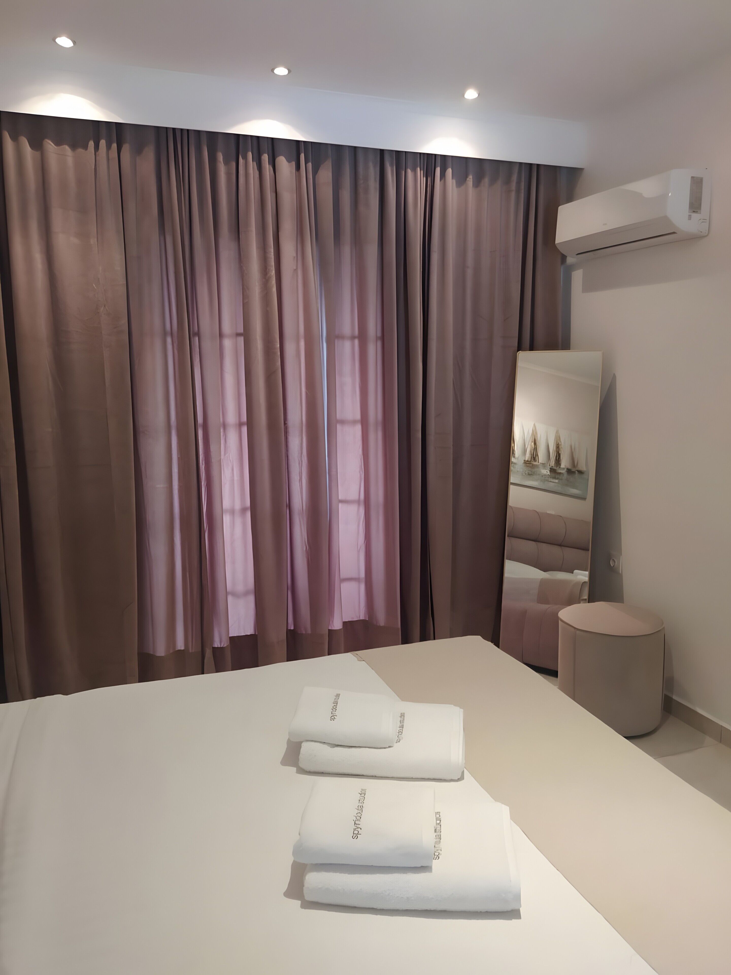 A hotel room with a large bed, a round bedside table, a wall-mounted air conditioner, and a mirror reflecting a bed and curtains.