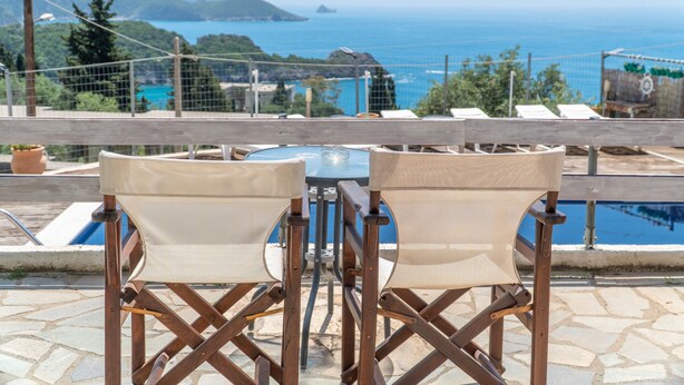 A terrace with a table and chairs, overlooking a pool and a scenic view of the sea and hills.