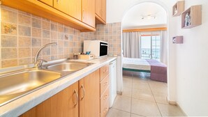A kitchen with a sink, microwave, and cabinets, leading to a bedroom with a bed and a window.