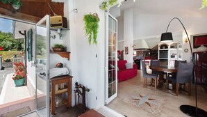 Une porte ouverte donne sur un espace de vie comprenant un canapé rouge, une table à manger et des chaises. Au fond, on aperçoit une cuisine, et un balcon agrémenté de plantes en pot.