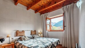 A bedroom with a wooden ceiling, a bed with a geometric patterned bedspread, a bedside table with a lamp, a window with curtains, and a view of mountains.