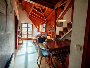 A spacious loft-style room with a high ceiling, wooden beams, a dining area with a table and chairs, a sofa, and a staircase.