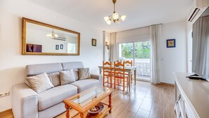 Un salon comprenant un canapé, une table basse, une table à manger avec des chaises, un lustre et une grande fenêtre avec des rideaux.