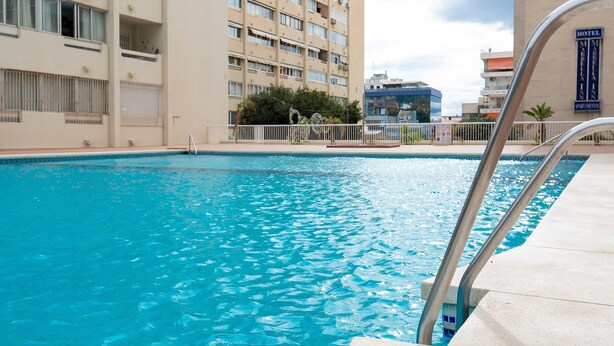 Une piscine avec un escalier en métal, entourée d’une clôture blanche et d’un bâtiment sur lequel est apposée une enseigne indiquant qu’il s’agit d’un hôtel.