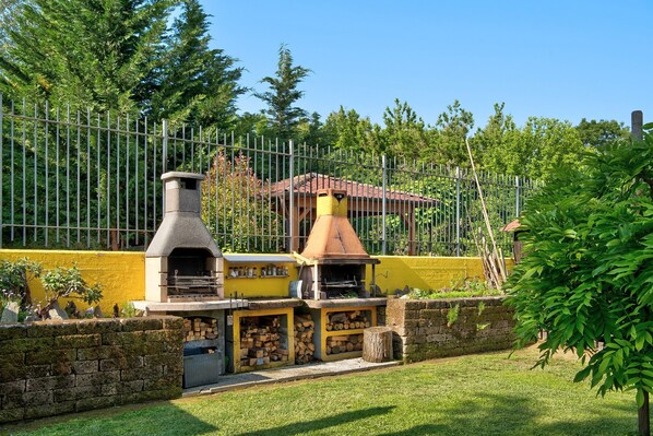 Un área de barbacoa con chimenea, horno de ladrillo y parrilla, rodeada por un muro amarillo y una valla metálica.