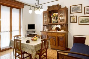 A dining area with a table set for four, a wooden cabinet with a vase of flowers, and a bed with a blue bedspread. - Holiday apartment 'Glicine' in Piedmont, with shared pool, garden and sauna (CISTERNA D'ASTI)