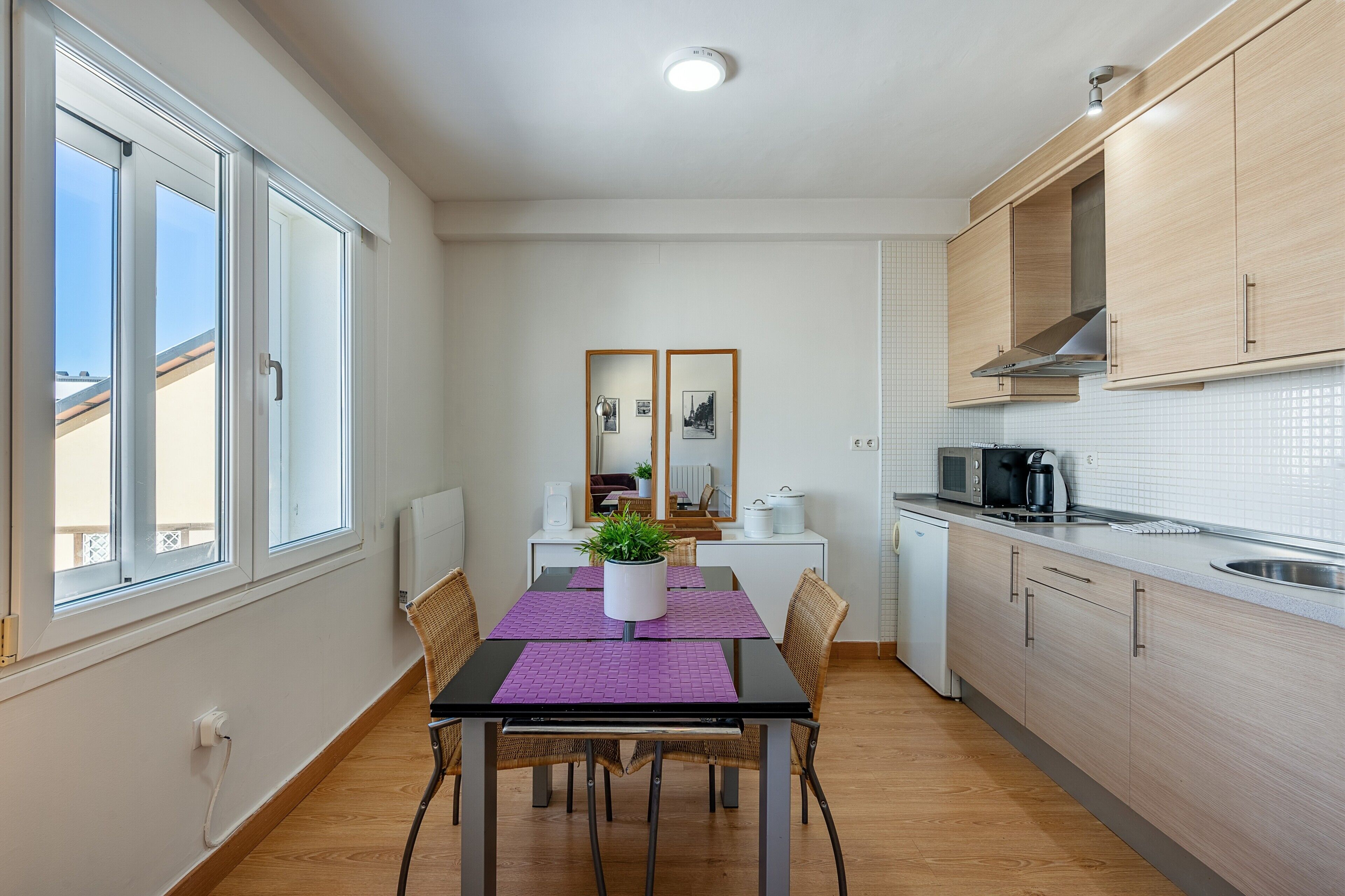 Een kleine keuken met een eettafel, stoelen, een magnetron, een koffiezetapparaat en een raam met jaloezieën.