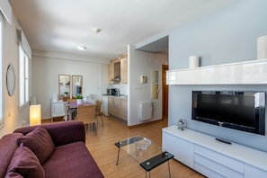 Una sala de estar moderna con un sofá, una mesa de centro de vidrio, un televisor de pantalla plana y una cocina al fondo.