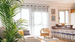 A living room with a sofa, a wicker chair, a small table with a 'Beach' sign, and a kitchen area with a bar stool.