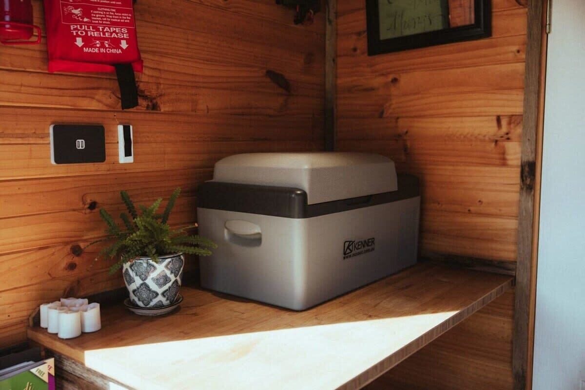 Una kitchenette compacta con un electrodoméstico Kenner, una pequeña planta en maceta y un dispensador de cinta roja.