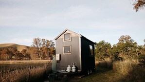 En liten, envåningsstuga med ett sluttande tak, en ytterdörr och en sidodörr. Den ligger på en gräsklädd äng med träd i bakgrunden.