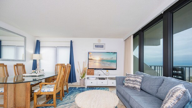 A modern living room with a glass dining table, wooden chairs, a grey sofa, and a large flat-screen TV. - Beach Front Condo with Bunks! (Gulf Shores)