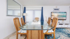 A dining area with a glass table surrounded by wooden chairs, a television on a white cabinet, and a 'Starfish' sign on the wall. - Beach Front Condo with Bunks! (Gulf Shores)