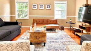 A living room with a sofa, coffee table, and TV. There are windows with curtains, a rug, and framed pictures on the wall.