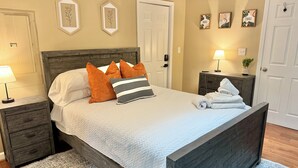 A bedroom with a wooden bed, white bedding, and orange pillows. There are framed pictures on the wall, a nightstand with a lamp, and a small table with a plant.