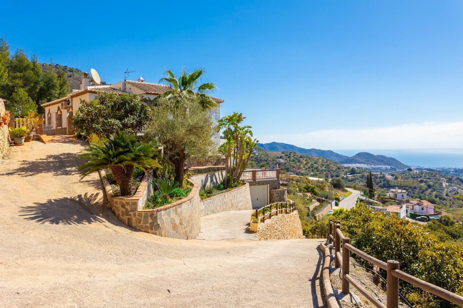 A hillside property with a stone pathway leading to a building, surrounded by lush greenery and a scenic view of the coastline.