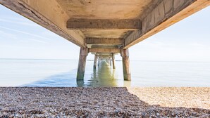 Un quai en béton s’étendant dans une étendue d’eau calme, avec une plage de galets au premier plan.