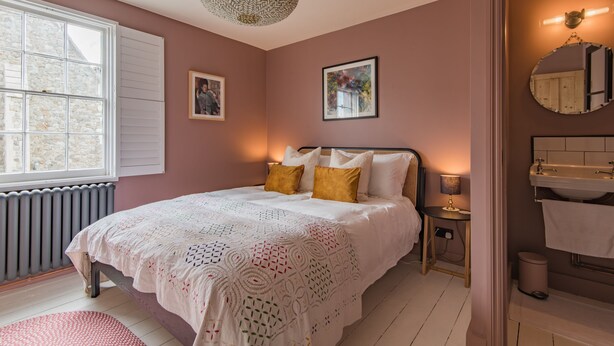 Une chambre avec un grand lit, une table de chevet avec une lampe, une fenêtre avec des volets et une salle de bain avec un lavabo et un miroir.