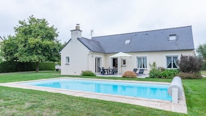 Une maison avec une piscine, une terrasse dotée de mobilier d’extérieur et un grand arbre dans le jardin.