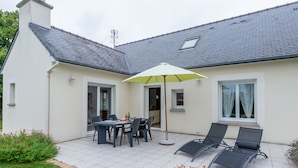 Une maison avec un patio, une table à manger avec des chaises, un parasol jaune et deux fauteuils de jardin.