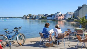 A waterfront café with two people seated at wicker tables, overlooking a calm harbor with boats and colorful buildings in the background.