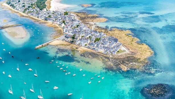 An aerial view of a coastal town with a prominent white building complex, a sandy beach, and clear turquoise waters with numerous boats.