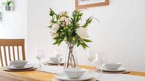 Una mesa de comedor dispuesta con platos, tazones y copas de vino blancos, y un florero con flores en el centro.
