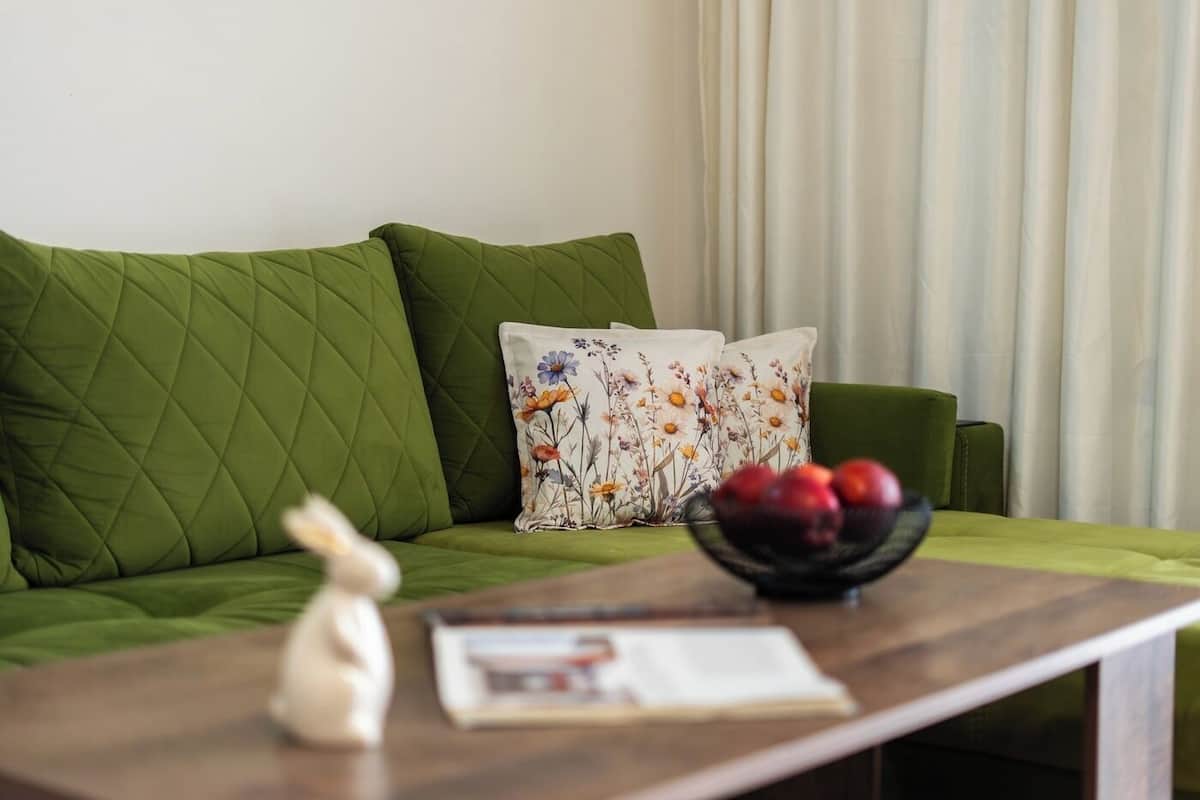 A green sofa with quilted cushions, a floral pillow, a small white figurine, a bowl of fruit, and a magazine on a wooden table.